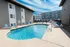 A bright outdoor swimming pool surrounded by lounge chairs and tables, adjacent to modern apartment buildings under a clear blue sky. Calm and inviting atmosphere.