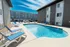 Outdoor pool surrounded by modern apartment buildings under a bright blue sky. Several lounge chairs with blue cushions line the poolside. Tranquil ambiance.