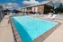 Outdoor swimming pool with clear blue water, surrounded by white lounge chairs on a sunny day. Brick building and parked cars in the background. Relaxing atmosphere.