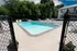 A serene outdoor swimming pool surrounded by a concrete deck, adjacent to a small parking lot and lush green trees under a partly cloudy sky.