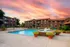 Beautiful courtyard view of an apartment complex at sunset, featuring a well-maintained swimming pool surrounded by lounge chairs, potted plants, and a small fountain in the foreground. The sky is painted with vibrant pink and orange hues, adding a warm ambiance to the scene