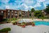 Daytime view of an apartment complex courtyard with a swimming pool, surrounded by white lounge chairs and potted plants. A small fountain is located in the foreground, with vibrant greenery and brick buildings in the background. The sky is clear with fluffy white clouds, creating a bright and inviting atmosphere.