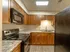 Compact kitchen with wooden cabinets, black appliances, and a marbled countertop. A stove, microwave, and double sink are visible, with overhead lighting providing ample illumination. The flooring has a wood-like finish, adding warmth to the space.