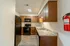 Galley-style kitchen with black appliances, including a refrigerator and stove, paired with wooden cabinets and a marbled countertop. A fire extinguisher is mounted on the wall, and the room is well-lit by a ceiling light. The wood-patterned floor complements the warm tones of the cabinetry.