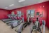 Quail Ridge Apartments fitness center with a row of treadmills, stationary bikes, and elliptical machines arranged along red walls. The space features large windows providing natural light and a carpeted floor for safe footing. Equipment is spaced to allow clear pathways for accessibility, ensuring an inclusive and accommodating workout environment for all residents.