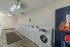 Quail Ridge Apartments community laundry room featuring rows of white washers and dryers, including a front-loading machine for accessibility. The space is well-lit with overhead lighting and ceiling fans, providing a clean and comfortable environment. A vending machine is visible on the right for added convenience.