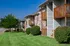 Side view of Grandview Apartments showcasing brick and siding buildings with private wooden balconies. The neatly trimmed bushes and lush green lawn provide a well-maintained appearance, with a clear blue sky and the Grandview Village sign visible in the background.