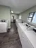 A view of a laundry room showing two rows of top-loading washing machines and dryers aligned neatly. The room is well-lit with overhead lighting, has gray tiled flooring, and light green walls. A window brings in some natural light, and an electrical panel is visible in the background.