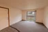A spacious living room with neutral-colored carpet and white walls. A sliding glass door leads to a private balcony with a wooden railing, offering a view of a grassy outdoor area.