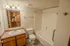 A clean bathroom featuring a white bathtub with built-in shelves and a grab bar, a single sink vanity with a wooden cabinet, a large mirror, and a toilet. A tripod is reflected in the mirror.