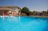 Outdoor swimming pool with clear blue water, surrounded by a black metal fence. Lounge chairs and a patio umbrella are placed on the pool deck. A two-story brick and siding apartment building is visible in the background under a bright blue sky.