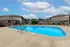 Outdoor swimming pool with clear blue water, surrounded by a concrete deck with metal handrails and depth markers. Lounge chairs and patio tables are arranged around the pool area, which is enclosed by a black metal fence. In the background, two-story brick apartment buildings with beige siding and a parking lot are visible under a partly cloudy sky. A sign on one of the buildings displays the property management company's name.
