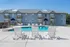 A three-story apartment building at Concord Manor with gray siding, brick accents, and wooden balconies. In the foreground, a fenced-in outdoor pool area features three white lounge chairs facing the water. Additional chairs and tables are placed around the pool deck. The sky is clear and blue, and the surrounding area includes trees and power lines in the background.