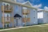 Another angle of the three-story Concord Manor apartment buildings, showcasing their uniform design with gray siding, brick accents, and wooden balconies. Some balconies have potted plants, and a well-maintained sidewalk runs along the front of the building under a partly cloudy sky.