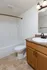 Bathroom featuring a white bathtub and shower combination with a curved shower rod, a toilet, and a vanity with a speckled brown countertop and a single sink. The vanity has light wood cabinetry with black hardware, a bronze faucet, and a mirror above with a two-light fixture. The floor is covered in beige and brown marbled flooring