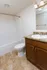 Bathroom featuring a white bathtub and shower combination with a curved shower rod, a toilet, and a vanity with a speckled brown countertop and a single sink. The vanity has dark wood cabinetry with silver hardware, a silver faucet, and a mirror above with a three-light fixture. The floor is covered in beige and brown marbled flooring