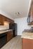 Galley-style kitchen with dark wood cabinetry, black appliances including a refrigerator, electric stove, microwave, and dishwasher. The speckled brown countertops contrast with the light wood-look vinyl flooring. Under-cabinet lighting illuminates the double sink area, and a flush-mounted ceiling light provides additional lighting.
