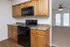 Kitchen with light wood cabinetry, black appliances including an electric stove and built-in microwave, and a speckled brown countertop. The space features wood-look flooring and ample counter space. In the background, a carpeted living area with a ceiling fan and French doors with built-in blinds is visible.