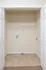 Open laundry closet at Embassy Ozark Apartments in Ozark, Missouri, with double white doors, a beige tiled floor, and beige walls. Inside, there is a single white wire shelf for storage and visible washer and dryer hookups, including hot and cold water connections and an exhaust outlet on the wall. The lower half of the floor transitions from tile to brown carpet just outside the closet opening.