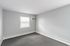 Bright and airy room with neutral gray walls and carpet, perfect for cozy living. Features a large window with blinds and built-in air conditioning unit.