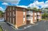 Charming brick apartment building with private balconies, nestled in a lush setting with ample parking under a bright blue sky.