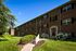 Charming brick apartment complex with lush green landscaping and a welcoming entrance, set against a clear blue sky.