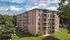 Charming brick apartment building surrounded by lush greenery and blue skies, featuring spacious balconies and serene surroundings.