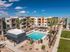 Modern apartment complex featuring a spacious swimming pool, lush palm trees, and stylish multi-level buildings under a bright blue sky.