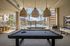 Luxurious apartment amenity area with a sleek pool table, foosball, and a stunning view of the sparkling pool and modern apartment buildings under a sunny sky.