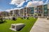Modern apartment complex with vibrant facade, lush green garden area, and outdoor seating under a clear blue sky.