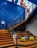 Stadium seating lounge in student housing Auburn AL. Modern multi-level interior featuring stylish wooden stairs, vibrant blue walls, and chic seating area with colorful ottomans, perfect for a trendy urban lifestyle.