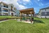 Charming outdoor picnic area with wooden pergola, surrounded by modern apartment complexes and lush green lawns under a bright blue sky.