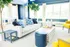 Bright and inviting living room with a modern white sofa, vibrant blue accents, and lush greenery framing large sunlit windows.