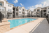 Modern apartment complex with a luxurious pool and spacious sunbathing area, set against a clear blue sky.