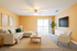 Bright and inviting living room with a modern beige sectional sofa, green accent pillows, and a wooden coffee table. Features a large flat-screen TV, a cozy armchair, and sliding doors opening to a sunlit balcony.