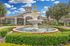Elegant community entrance with a grand fountain in a lush landscaped setting, showcasing classic architecture under a bright blue sky.