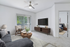 Modern living room with plush gray sofa, wooden coffee table, and wall-mounted TV, adjacent to a cozy bedroom. Natural light floods through large patio doors, creating a bright, inviting space.