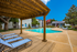Inviting outdoor pool area with sun loungers under a wooden pergola, surrounded by lush greenery and charming buildings.