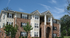 Charming three-story brick apartment building with large windows, elegant columns, and lush green landscaping under a clear blue sky.