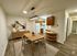 Inviting dining area with a modern light fixture, elegant wood table, and chairs adjacent to a cozy kitchen with warm wooden cabinets and appliances. Perfect for intimate gatherings and culinary creativity.