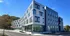 Modern four-story apartment building with sleek glass windows, surrounded by lush greenery and a clear blue sky.