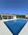 Inviting outdoor swimming pool with pristine blue water, sun loungers, and a covered seating area, surrounded by lush greenery and clear skies.