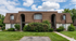 Charming two-story apartment building with a classic brown shingle roof, featuring lush greenery and a welcoming pathway against a bright blue sky.