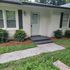 Charming front entrance of a cozy home with a welcoming teal door, surrounded by lush greenery and neatly trimmed bushes.