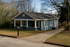 Charming single-story blue house with a cozy front porch and well-maintained lawn, nestled among tall, mature trees.