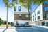 Modern apartment complex exterior with contemporary design and large windows, surrounded by lush greenery and sunny skies.