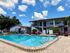 Charming apartment complex with a sparkling pool surrounded by sun loungers, vibrant umbrellas, and lush greenery under a bright blue sky.