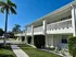Charming two-story apartment building with white railings, surrounded by lush greenery and palm trees under a bright blue sky.