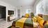 Bright and modern bedroom featuring a stylish bed with vibrant yellow and green bedding, complemented by a cozy armchair and elegant chest of drawers beneath a sleek wall-mounted TV.