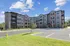Modern four-story apartment building with brick and stucco facade, surrounded by lush landscaping and ample parking, under a clear blue sky.